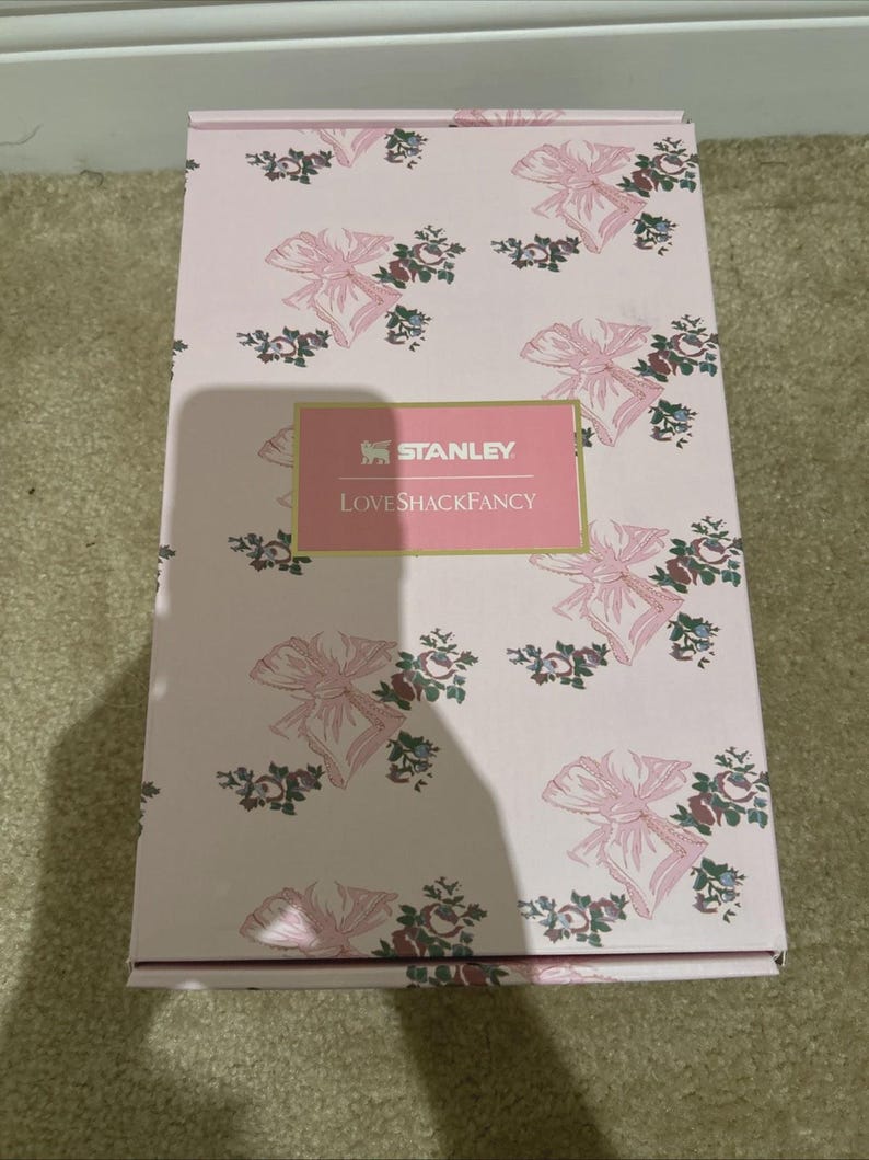 Puede incluir: Una caja rosa con un estampado de lazos y flores. La caja tiene el texto "STANLEY" y "LoveShackFancy" en una etiqueta rosa. La caja es rectangular y parece nueva.