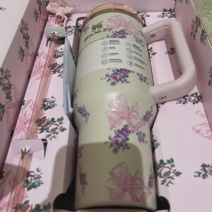 Puede incluir: Un vaso Stanley Quencher en una caja de regalo. El vaso crema presenta lazos rosas y estampados florales morados. Tiene un asa y tapa rosas. La caja de regalo es rosa con un dise&ntilde;o floral. Se incluye una pajita rosa.
