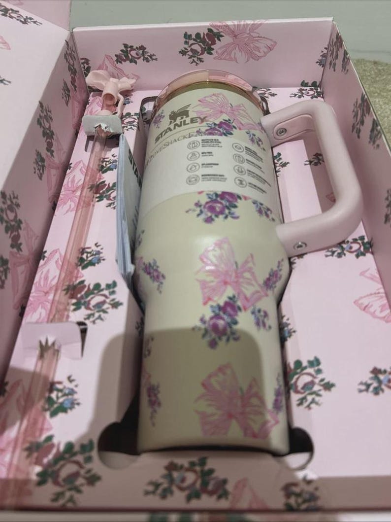 Puede incluir: Un vaso Stanley Quencher en una caja de regalo. El vaso crema presenta lazos rosas y estampados florales morados. Tiene un asa y tapa rosas. La caja de regalo es rosa con un dise&ntilde;o floral. Se incluye una pajita rosa.