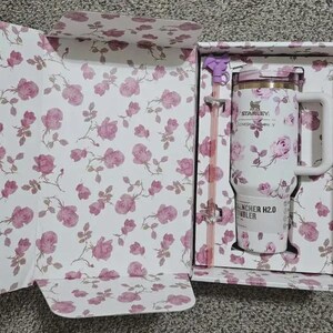 May include: A white Stanley Quencher H2.0 tumbler with a floral rose pattern, presented in a matching floral box. The tumbler has a handle and straw, and the box is open, revealing the design. The overall color scheme is white and pink.