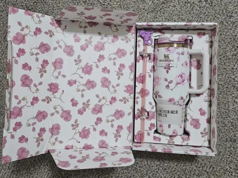 May include: A white Stanley Quencher H2.0 tumbler with a floral rose pattern, presented in a matching floral box. The tumbler has a handle and straw, and the box is open, revealing the design. The overall color scheme is white and pink.