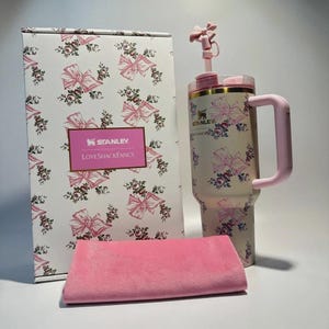 Puede incluir: Un vaso Stanley LoveShackFancy con asa y pajita rosas, una toalla rosa a juego y una caja. El vaso y la caja presentan un fondo blanco con lazos rosas y motivos florales. La caja tiene los logotipos de Stanley y LoveShackFancy.