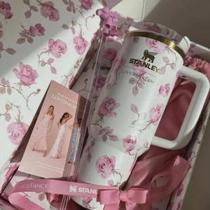 May include: A white Stanley tumbler with a pink rose pattern, inside a gift box with matching floral design. The box includes a pink ribbon, a card with images of dresses, and the text "LOVESHACKFANCY".
