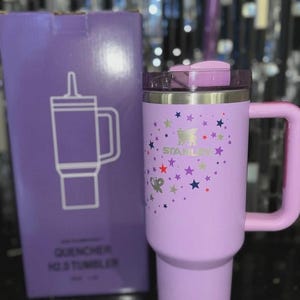 May include: A lavender Stanley Quencher H2.0 tumbler with a handle and lid, decorated with colorful stars. The tumbler is next to a matching box with a line drawing of the tumbler and the words "QUENCHER H2.0 TUMBLER".