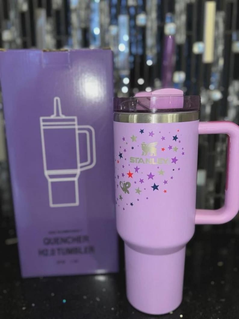 May include: A lavender Stanley Quencher H2.0 tumbler with a handle and lid, decorated with colorful stars. The tumbler is next to a matching box with a line drawing of the tumbler and the words "QUENCHER H2.0 TUMBLER".