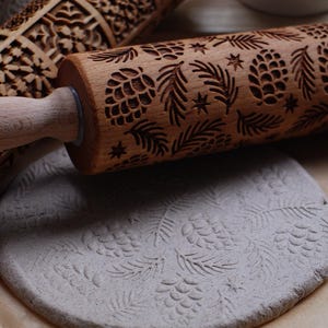 Puede incluir: Rodillos de madera con intrincados diseños tallados, incluyendo piñas, ramas de abeto y estrellas. Un rodillo está rodando sobre una masa gris, creando un patrón en relieve a juego. Los rodillos son de madera marrón claro.