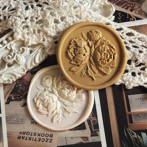May include: Two circular wax seals featuring a raised floral peony design. One seal is a solid tan color, while the other is a creamy white. Both seals have a smooth, raised border and detailed, embossed flower petals and leaves on their surfaces.