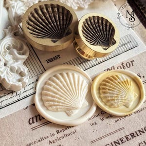 Peut inclure: Deux sceaux en laiton circulaires gravés d'un coquillage, accompagnés de deux cachets de cire correspondants. L'un est blanc cassé uni, l'autre présente un mélange marbré de crème et d'or, tous deux reproduisant le motif du coquillage.