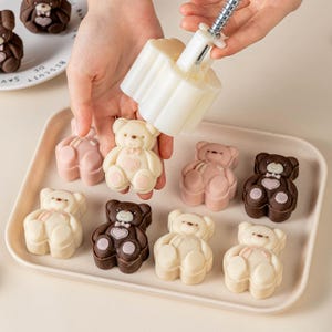 Può includere: Una persona usa uno stampo bianco per creare dolcetti a forma di orsetto. I dolcetti sono in vari colori, tra cui bianco, rosa e marrone, e sono disposti su un vassoio bianco. I dolcetti hanno dettagli a forma di cuore.