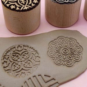 May include: Close-up of wooden stamps with floral and geometric designs. Two stamps are visible, along with impressions on a clay surface. The stamps have a natural wood finish, and the impressions are in a light brown color.