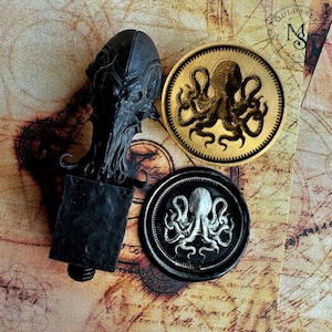 Puede incluir: Un sello de cera negro con un mango detallado en forma de cabeza inspirada en Cthulhu, un sello de cera circular dorado con un diseño de pulpo y un sello de cera circular negro con un emblema de pulpo plateado sobre un fondo de mapa.