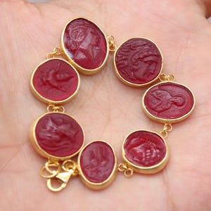 Puede incluir: Un brazalete dorado con siete cabujones ovalados de piedras preciosas rojas, cada uno con un diseño en relieve tallado. Las piedras están engastadas en biseles dorados y unidas con pequeños conectores dorados.