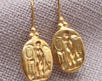 May include: A pair of gold-colored earrings with an oval pendant featuring a detailed relief of a figure. The earrings have a hook closure and a small beaded accent above the pendant. The background is a neutral fabric.