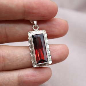 May include: A rectangular pendant with a large, deep red gemstone. The gem is set in a silver-colored, textured frame. The pendant is suspended from a small loop at the top.