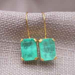 Puede incluir: Pendientes dorados con piedras preciosas rectangulares de color verde esmeralda. Los pendientes tienen un diseño sencillo con cierre de gancho. Las piedras están engastadas en un bisel dorado, creando un aspecto clásico y sofisticado.