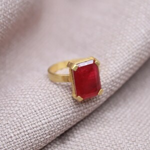 Peut inclure: Une bague dor&eacute;e avec une grosse pierre pr&eacute;cieuse rectangulaire rouge fonc&eacute;. La bague est pos&eacute;e sur un tissu textur&eacute; de couleur claire. La pierre pr&eacute;cieuse est le point central, captant la lumi&egrave;re.