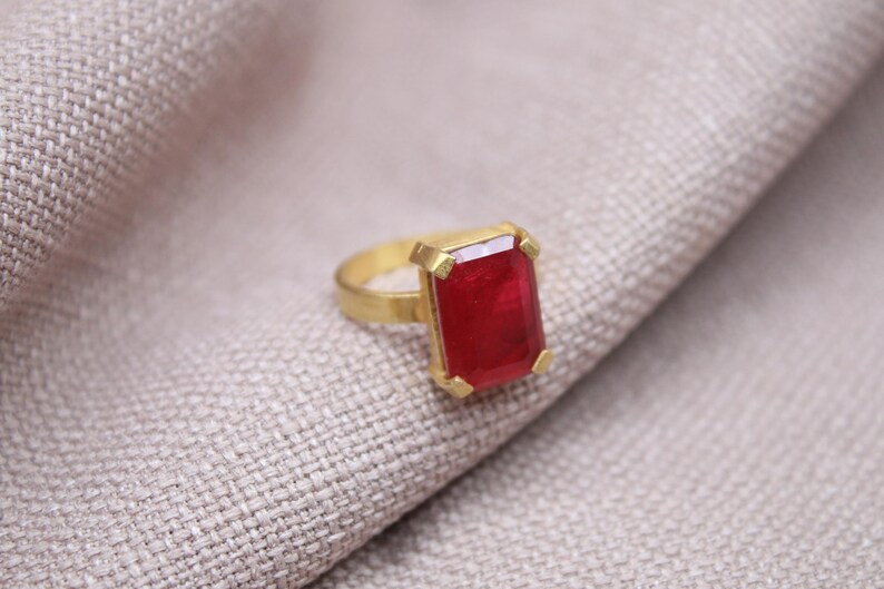 Peut inclure: Une bague dor&eacute;e avec une grosse pierre pr&eacute;cieuse rectangulaire rouge fonc&eacute;. La bague est pos&eacute;e sur un tissu textur&eacute; de couleur claire. La pierre pr&eacute;cieuse est le point central, captant la lumi&egrave;re.