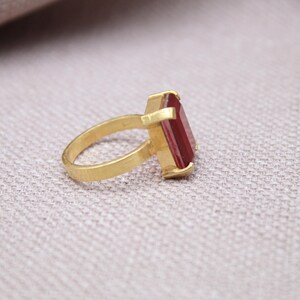 Peut inclure: Une bague en or avec une pierre pr&eacute;cieuse rectangulaire rouge fonc&eacute;. La bague est pos&eacute;e sur une surface claire, la monture et le corps en or contrastant avec la pierre rouge. La bague est inclin&eacute;e, mettant en valeur son design.