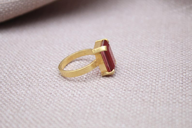 Peut inclure: Une bague en or avec une pierre pr&eacute;cieuse rectangulaire rouge fonc&eacute;. La bague est pos&eacute;e sur une surface claire, la monture et le corps en or contrastant avec la pierre rouge. La bague est inclin&eacute;e, mettant en valeur son design.