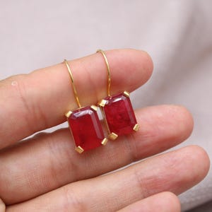 May include: A pair of gold drop earrings featuring rectangular, deep red gemstones. The earrings are held in a hand against a neutral background. The gemstones have a faceted appearance.
