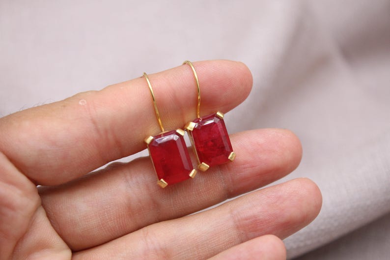May include: A pair of gold drop earrings featuring rectangular, deep red gemstones. The earrings are held in a hand against a neutral background. The gemstones have a faceted appearance.