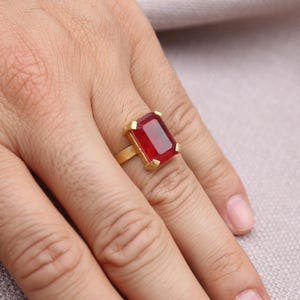 Peut inclure: Une bague dor&eacute;e orn&eacute;e d'une grande pierre rouge rectangulaire. La bague est port&eacute;e au doigt, mettant en valeur le design et la couleur du bijou. Le sertissage de la bague est simple et &eacute;l&eacute;gant.