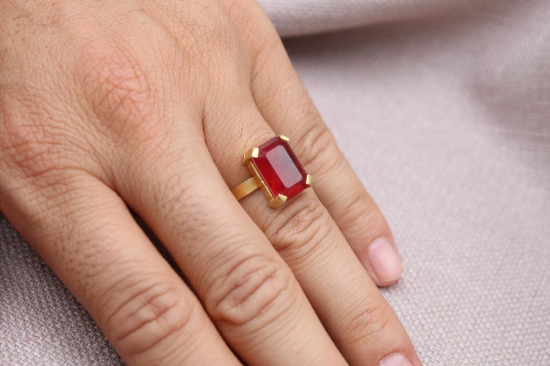 Peut inclure: Une bague dor&eacute;e orn&eacute;e d'une grande pierre rouge rectangulaire. La bague est port&eacute;e au doigt, mettant en valeur le design et la couleur du bijou. Le sertissage de la bague est simple et &eacute;l&eacute;gant.