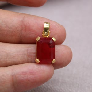 May include: A rectangular, faceted, deep red gemstone pendant with a gold-colored bail and prongs. The pendant is held in a hand, showcasing its vibrant color and design. The gemstone is approximately 0.75 inches in length.
