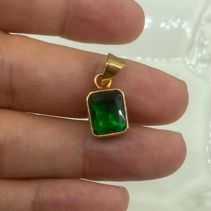 May include: A gold pendant with a rectangular, emerald-green gemstone. The pendant has a simple, elegant design with a gold bail for a chain. The gemstone is faceted, catching the light and displaying a rich, deep green color.
