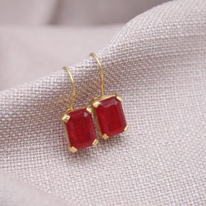 May include: Gold-coloured earrings featuring rectangular, deep red gemstones. The earrings are set in a gold frame and hang from a simple gold hook. The jewellery rests on a textured, light grey fabric.