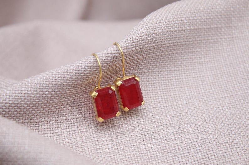 May include: Gold-coloured earrings featuring rectangular, deep red gemstones. The earrings are set in a gold frame and hang from a simple gold hook. The jewellery rests on a textured, light grey fabric.