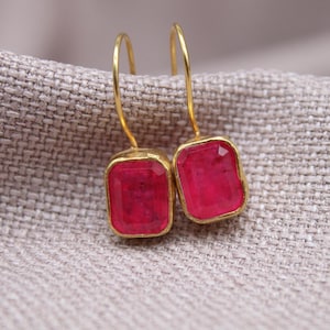May include: A pair of gold-colored earrings with rectangular, faceted, bright pink gemstones. The earrings are displayed on a textured, light gray fabric. The gemstones have a vibrant, eye-catching color.