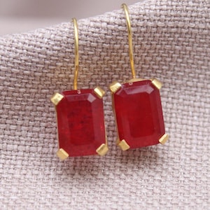 May include: A pair of rectangular red gemstone earrings with gold-colored accents. The earrings have a gold-colored hook and are set against a neutral fabric background. The gemstones are a deep red color.