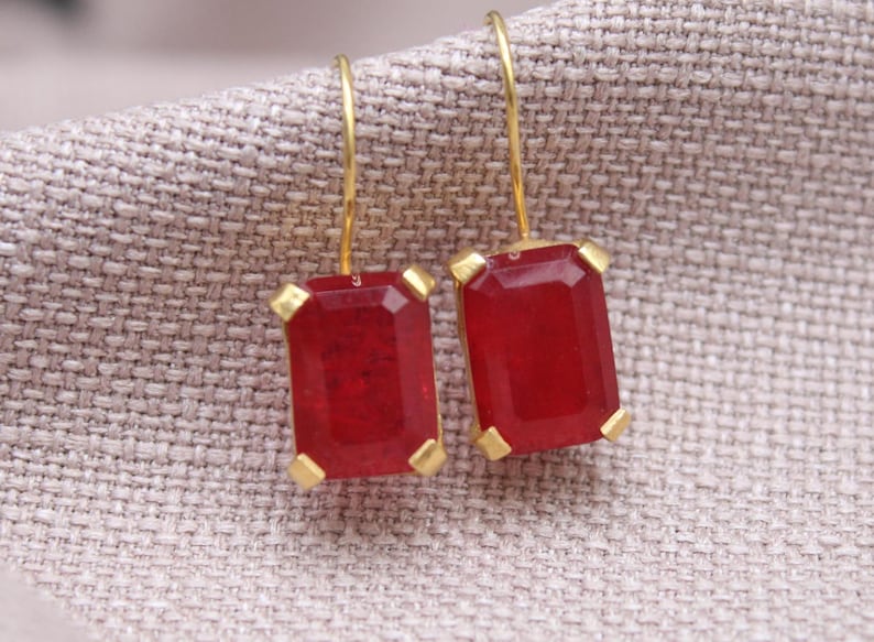 May include: Earrings featuring rectangular, deep red gemstones with gold-coloured settings and hooks. The earrings are displayed against a neutral fabric backdrop. The gemstones are a rich, red hue.