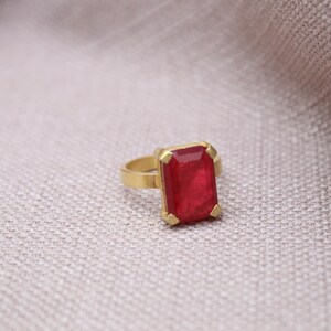 Peut inclure: Une bague dor&eacute;e orn&eacute;e d'une grande pierre rouge rectangulaire. La pierre est sertie dans un sertissage en or. La bague est pr&eacute;sent&eacute;e sur une surface neutre et textur&eacute;e.