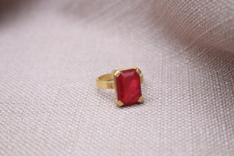Peut inclure: Une bague dor&eacute;e orn&eacute;e d'une grande pierre rouge rectangulaire. La pierre est sertie dans un sertissage en or. La bague est pr&eacute;sent&eacute;e sur une surface neutre et textur&eacute;e.