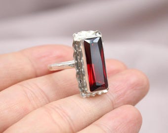 May include: A silver ring featuring a rectangular, deep red gemstone. The gemstone is set within a textured silver bezel. The ring is held in a hand, highlighting the design and colour of the jewellery.