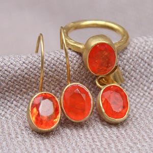 May include: A jewelry set featuring a gold ring and earrings with oval, orange gemstones. The ring has a single gemstone, while the earrings have two. The jewelry is displayed on a textured, neutral-colored surface.