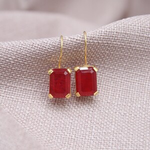 May include: Gold-coloured drop earrings featuring rectangular, deep red gemstones. The earrings are set in gold and suspended from fine gold wires. The background is a neutral, textured fabric.