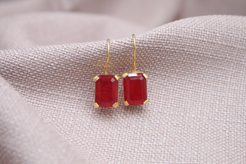 May include: Gold-coloured drop earrings featuring rectangular, deep red gemstones. The earrings are set in gold and suspended from fine gold wires. The background is a neutral, textured fabric.