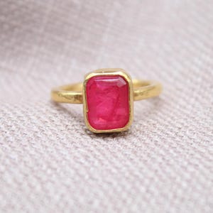 May include: A gold ring with a rectangular, vibrant pink gemstone. The ring's band and setting are a warm gold color, creating a striking contrast with the gemstone. The ring is set against a neutral, textured background.