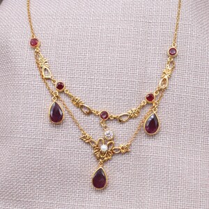 Puede incluir: Un collar dorado con detalles de rubíes y piedras preciosas transparentes. El collar presenta un diseño floral con colgantes de rubí en forma de lágrima y pequeños rubíes redondos. El collar se muestra sobre una superficie de color neutro.