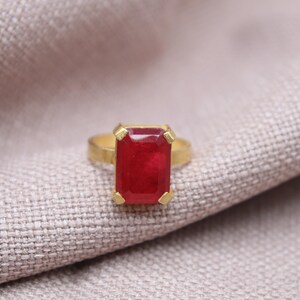 Peut inclure: Une bague en or avec une grosse pierre rouge rectangulaire. La bague est pos&eacute;e sur un tissu clair et textur&eacute;. Le rouge vif de la pierre attire l'attention.