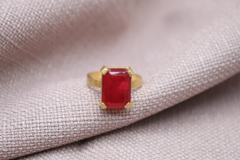 Peut inclure: Une bague en or avec une grosse pierre rouge rectangulaire. La bague est pos&eacute;e sur un tissu clair et textur&eacute;. Le rouge vif de la pierre attire l'attention.