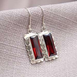 May include: A pair of silver earrings with rectangular, deep red gemstones. The gemstones are set in a textured, silver frame with a hook closure. The earrings are displayed on a neutral-colored fabric.