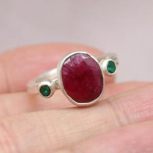 May include: A silver ring featuring a large, oval-shaped, deep red ruby gemstone set in the center, flanked by two smaller, round, vibrant green emerald gemstones on either side. The metal band has a smooth, matte finish and a simple, clean design.