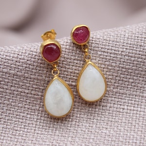 May include: A pair of gold-toned drop earrings. Each earring features a small, round, deep red gemstone at the top, connected to a teardrop-shaped, white gemstone. The gemstones are set in gold-colored metal.