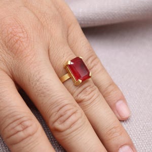 Peut inclure: Une bague en or avec une grosse pierre rouge rectangulaire. La bague est sertie dans un anneau en or et est pr&eacute;sent&eacute;e sur un doigt. La pierre pr&eacute;cieuse est d'un rouge profond et est le point central de la bague.
