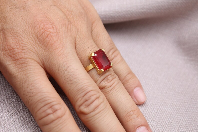 Peut inclure: Une bague en or avec une grosse pierre rouge rectangulaire. La bague est sertie dans un anneau en or et est pr&eacute;sent&eacute;e sur un doigt. La pierre pr&eacute;cieuse est d'un rouge profond et est le point central de la bague.
