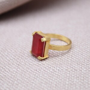 Peut inclure: Une bague dor&eacute;e avec une pierre pr&eacute;cieuse rectangulaire rouge fonc&eacute;. Le corps et le sertissage de la bague ont une finition mate. La pierre est sertie dans un sertissage &agrave; quatre griffes, sur une surface textur&eacute;e et claire.